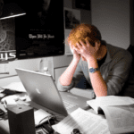A fatigued man sits at his desk, head in hand, surrounded by papers and a glowing screen, showing signs of burnout.