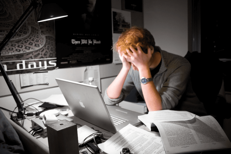 A fatigued man sits at his desk, head in hand, surrounded by papers and a glowing screen, showing signs of burnout.
